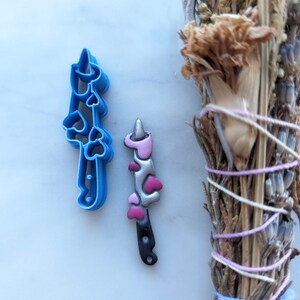 Peut inclure: Un emporte-pièce en plastique bleu et un objet décoré en forme de couteau sur le thème de la licorne. Le couteau a un manche noir, des détails roses et argentés et des accents en forme de cœur. Herbes séchées en arrière-plan.