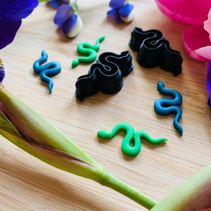 May include: Four small snake-shaped polymer clay charms in green, blue, and black. The charms are on a wooden surface with flowers in the background.