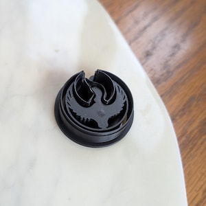 May include: A black plastic cookie cutter in the shape of a bird with outstretched wings, set on a white marble surface. The cutter has a circular outer edge and a detailed interior design.