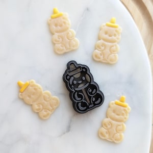 May include: A collection of bear-shaped cookie cutters and cookies. The cookies are light beige with yellow hats, and the cutter is black. The items are arranged on a white marble surface with a wooden border.