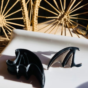 May include: Two black bat-shaped cookie cutters on a white surface. The cutters are made of plastic and have a shiny finish.