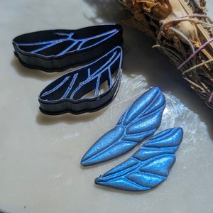 May include: Two blue iridescent resin fairy wings with a detailed vein pattern. The wings are next to two black plastic moulds of the same design.