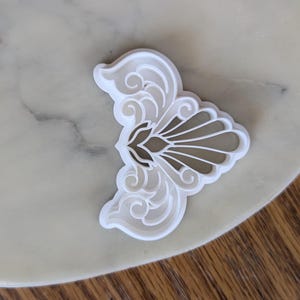 May include: A white plastic cookie cutter in the shape of a stylized butterfly or floral design. The cutter features intricate details with curved lines and a symmetrical pattern. The cutter is on a light-colored surface.