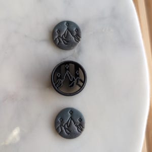 Könnte beinhalten: Drei schwarze Polymer Clay-Knöpfe mit einem Bergketten-Design. Die Knöpfe sind rund und haben ein erhabenes Design. Die Knöpfe befinden sich auf einer weißen Marmorfläche.