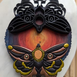 May include: A black and gold moth cookie cutter with a detailed design. The cutter is on top of a painted wooden round with a sunset design.