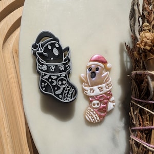 May include: Two Christmas stocking ornaments, one black with white details and one pink with white details. Both ornaments feature a ghost wearing a Santa hat.
