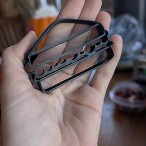 Puede incluir: Un cortador de galletas de plástico negro con forma de sándwich. El cortador tiene forma rectangular con bordes ondulados para representar los rellenos del sándwich. El cortador se sostiene en una mano.