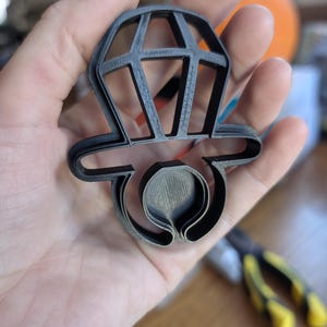 Puede incluir: Un cortador de galletas de plástico gris oscuro con forma de anillo de compromiso de diamantes. El cortador tiene un diseño detallado con forma de diamante y una banda de anillo. El cortador se sostiene en una mano.