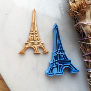 May include: Two Eiffel Tower cookie cutters, one gold and one blue. The gold cutter is solid, while the blue cutter is an outline.