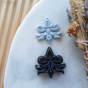May include: Two metal cookie cutters in the shape of a stylized flower. The top cutter is silver and the bottom cutter is black.