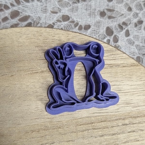 May include: A lavender-colored cookie cutter in the shape of a frog. The cutter is made of plastic and has a detailed design, including the frog's eyes, legs, and body. The cutter is on a wooden surface.
