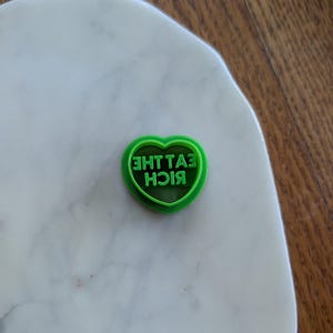 May include: A green heart-shaped cookie cutter with the words "EAT THE RICH" in reverse. The cutter is on a white marble surface, with a wooden background.