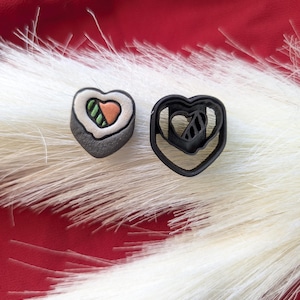 Puede incluir: Un trozo de sushi en forma de corazón y un cortador de galletas negro en forma de corazón. El sushi tiene un exterior gris, arroz blanco y rellenos naranjas y verdes. El cortador es de plástico negro. Los artículos están sobre una superficie blanca y esponjosa con un fondo rojo.