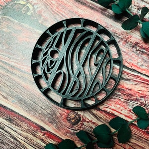 May include: Black circular coaster with a decorative design of intricate lines and curves. The coaster is made of a durable material and has a smooth finish.
