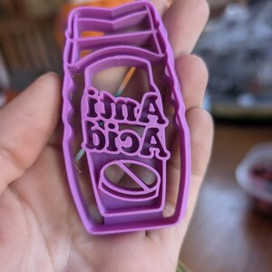 May include: A purple plastic cookie cutter in the shape of a bottle of antacid with the words "Anti Acid" in a stylized font. The cutter also features a pill shape inside the bottle.