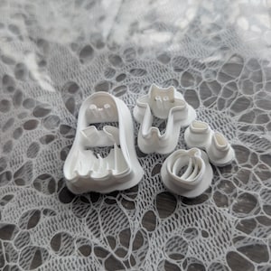 May include: A set of four white plastic cookie cutters featuring a ghost shape, a cat shape, a pumpkin shape, and a small pair of boots shape. Each cutter has a raised edge for cutting dough and a base for stability during the baking process.