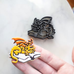 Puede incluir: Dos pines de esmalte que representan un dragón amarillo con alas naranjas durmiendo sobre un libro. Un pin es un contorno negro del dragón y el libro.