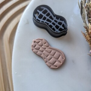 以下が含まれることがあります： 黒いピーナッツ型のクッキーカッターと、明るい茶色のピーナッツ型の粘土片。 クッキーカッターは格子模様で、粘土片はテクスチャ加工された表面です。 白い表面に置かれたアイテム。