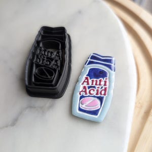 May include: Two black cookie cutters, one shaped like a bottle of pills with the text "Anti Acid" and the other shaped like a bottle of pills with the text "Mama Drama".