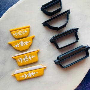 May include: A set of four yellow cookie cutters with white floral designs and four black cookie cutters in the shapes of bowls and casserole dishes.