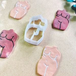 Puede incluir: Cuatro cortadores de galletas con forma de cuerpo humano, uno de plástico blanco y tres de arcilla rosa y marrón. Los cortadores están sobre una superficie marrón.