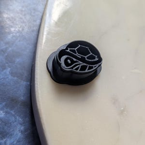 Peut inclure: Objet noir en forme de tortue avec un motif de carapace détaillé. L'objet est probablement un élément décoratif ou un composant d'un produit plus grand. La tortue est placée sur une surface claire, créant un contraste avec sa couleur sombre.