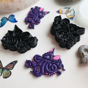 May include: Two purple and pink octopus cat cookie cutters, one with a detailed design and the other with a simpler design. The cutters are on a white surface with butterflies and a piece of a geode.