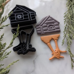 Puede incluir: Dos cortadores de galletas con forma de casa con patas. La casa es marrón y tiene un solo ojo en la parte delantera. Las patas son de color naranja. Los cortadores de galletas están sobre una superficie de mármol blanco.