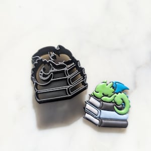 Puede incluir: Un cortador de galletas negro con forma de dragón enroscado en una pila de libros. También se muestra una galleta de dragón verde y azul, enroscada en una pila de libros.