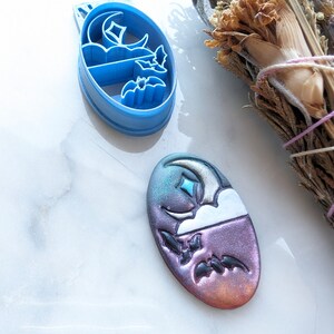 May include: A blue plastic cookie cutter with an oval shape, featuring a crescent moon, star, cloud, and two bats. Next to it is a finished oval-shaped piece with a gradient of purple, blue, and orange, with the same design.