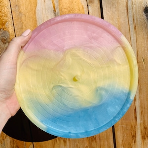 Puede incluir: Una bandeja redonda de resina con una combinación de colores pastel. La bandeja está dividida en tres secciones: rosa, amarillo y azul. Tiene un borde elevado y un elemento cilíndrico central. La bandeja es sostenida por una mano. Diámetro aproximado de 15 cm.