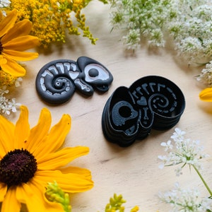 May include: Two black chameleon-shaped cookie cutters, one with white bone detailing and the other a solid outline. The cutters are on a light wood surface, surrounded by yellow and white wildflowers. The design is whimsical and suitable for baking.