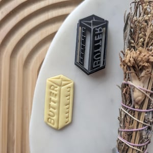 May include: A light yellow butter-shaped cookie cutter and a black cookie cutter with the word "BOLLER" printed on it. The butter cutter has the word "BUTTER" printed on it. The items are on a white surface.