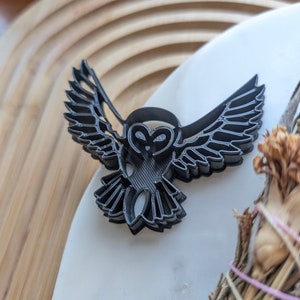 May include: A black plastic owl-shaped cookie cutter with detailed wing and feather patterns. The owl is shown with outstretched wings. The cutter is on a white surface, with a blurred wooden background.