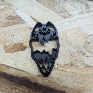 May include: A black plastic cookie cutter shaped like a melting ice cream cone. The design features a top scoop with a cat-like face, a middle section with dripping edges, and a bottom cone section with a cross-hatched grid pattern throughout.