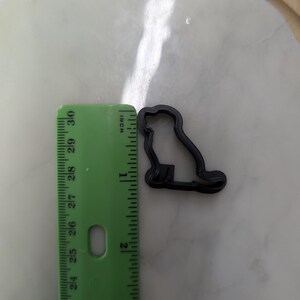 Puede incluir: Cortador de galletas de plástico negro con forma de perro. El cortador mide aproximadamente 5 cm de largo. Una regla verde con medidas en pulgadas está al lado del cortador. El fondo es una superficie de color claro.