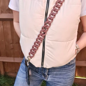 May include: A person wearing a white puffer vest with a brown chain phone strap. The strap is attached to a black phone case. The person is wearing blue jeans.