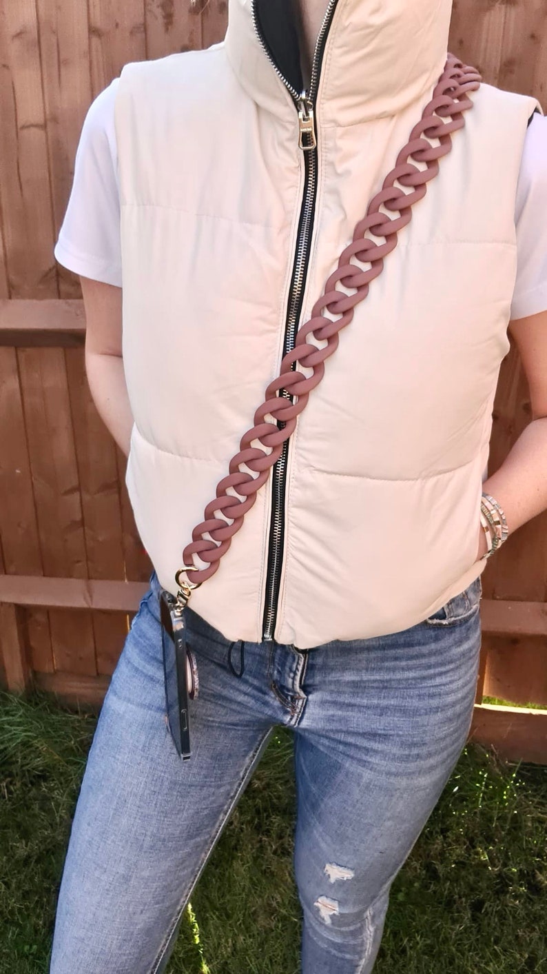 May include: A person wearing a white puffer vest with a brown chain phone strap. The strap is attached to a black phone case. The person is wearing blue jeans.