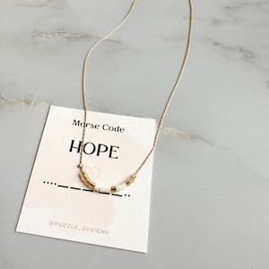 Puede incluir: Collar de cadena dorada con cuentas blancas que deletrean la palabra "HOPE" en código Morse. El collar se muestra en una tarjeta blanca con la palabra "HOPE" y el código Morse de la palabra.