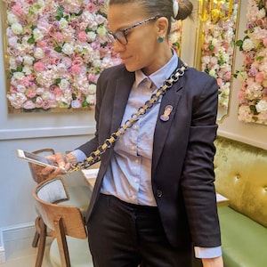 May include: A woman wearing a black blazer, a light blue button-down shirt, and black pants. She is holding a smartphone in her hand and has a gold chain with a small pin attached to it. The chain is worn as a crossbody strap.