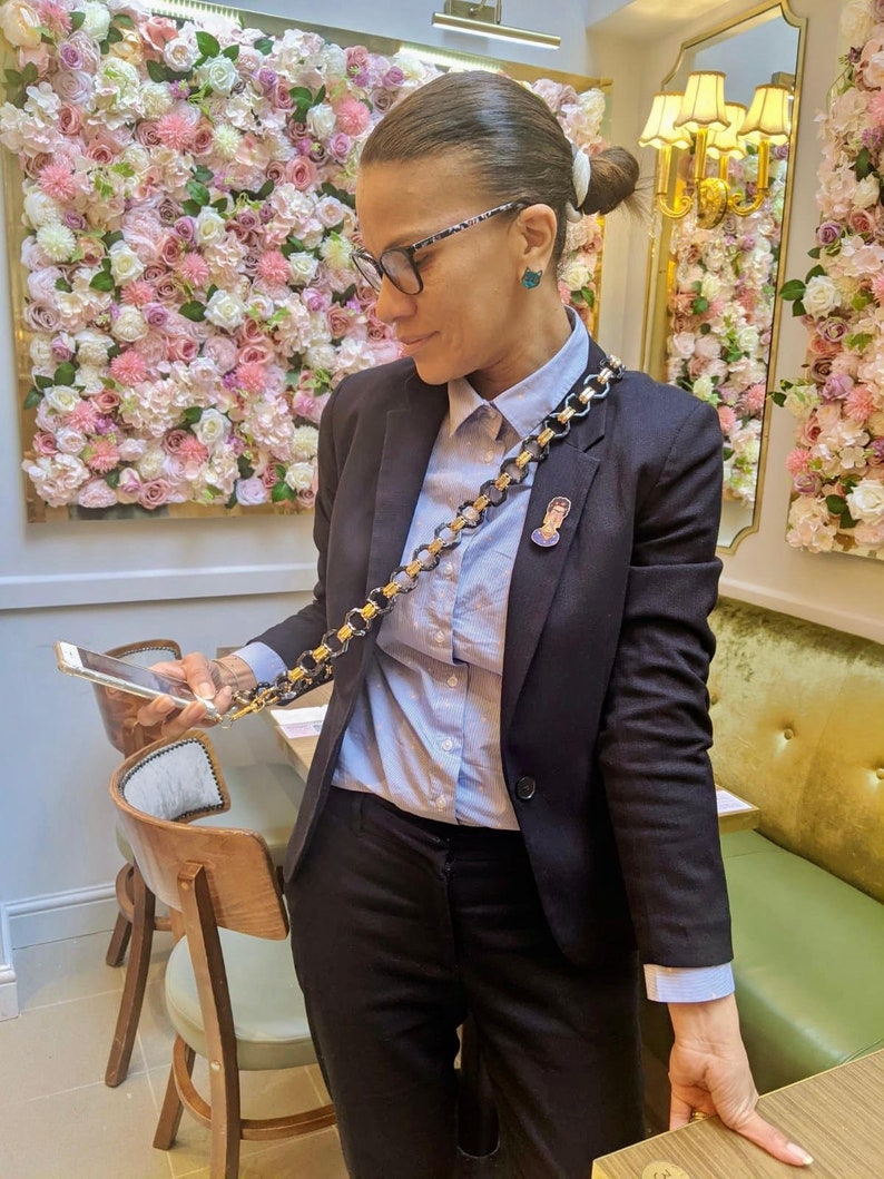 May include: A woman wearing a black blazer, a light blue button-down shirt, and black pants. She is holding a smartphone in her hand and has a gold chain with a small pin attached to it. The chain is worn as a crossbody strap.