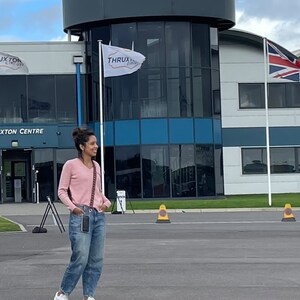 May include: A woman wearing a pink sweater and blue jeans stands in front of a building with a sign that reads "Thruxton Circuit".