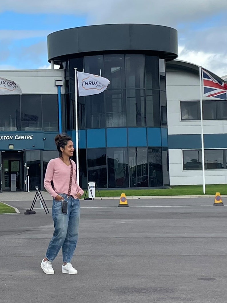 May include: A woman wearing a pink sweater and blue jeans stands in front of a building with a sign that reads "Thruxton Circuit".