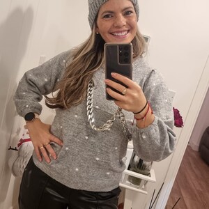 May include: A woman wearing a gray knitted sweater with white polka dots, black leather pants, and a gray knitted beanie. She is holding a smartphone in front of her face and smiling.
