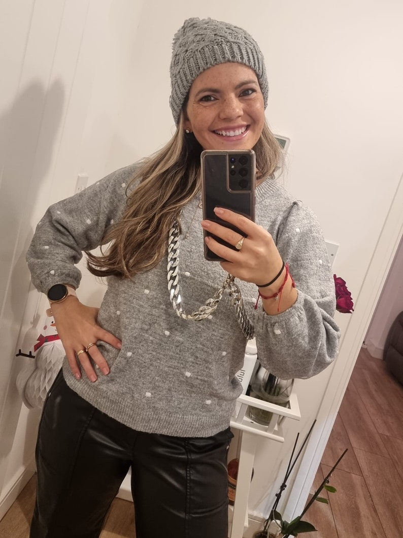 May include: A woman wearing a gray knitted sweater with white polka dots, black leather pants, and a gray knitted beanie. She is holding a smartphone in front of her face and smiling.