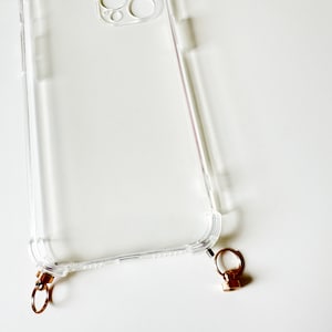 May include: A clear plastic phone case with a gold metal ring attached to the bottom. The case has a raised lip around the camera lens.