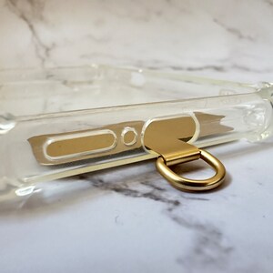 May include: A clear phone case with a gold metal ring attached to the bottom. The ring is designed to be used as a carrying handle or to attach a strap.