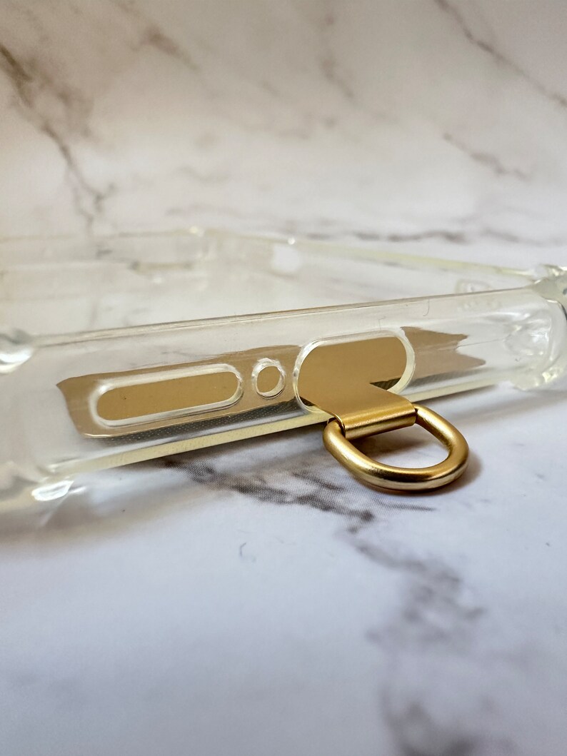 May include: A clear phone case with a gold metal ring attached to the bottom. The ring is designed to be used as a carrying handle or to attach a strap.