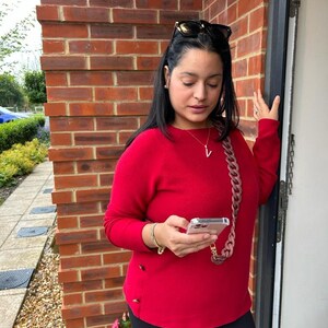 May include: A woman wearing a red sweater, black pants, and sunglasses is holding a smartphone with a pink chain strap. She is standing in front of a brick wall.