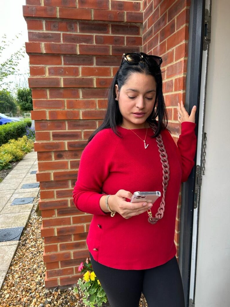 May include: A woman wearing a red sweater, black pants, and sunglasses is holding a smartphone with a pink chain strap. She is standing in front of a brick wall.